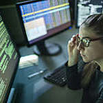 A young woman is seated at a desk surrounded by monitors displaying data, she is contemplating in this dark, moody office.